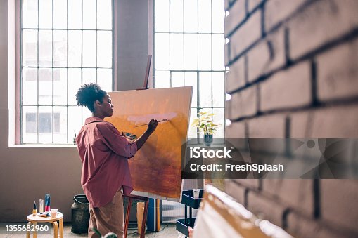 istock Beautiful Afro Female Artist Wearing a Pink Shirt, Painting on Canvas in her Home Studio 1355682960