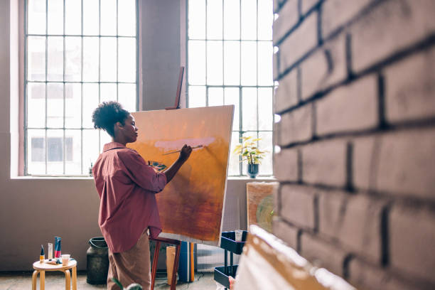 красивая афро-художница в розовой рубашке, рисуя на холсте в своей домашней студии - painted image painter women art стоковые фото и изображения