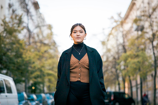 Portrait of a confident young woman standing outside. Asian female wearing fashionable clothing on city street and looking at camera.