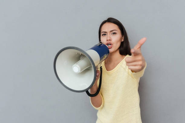 istock Woman in sweater screaming in megaphone and pointing at camera 1355687394