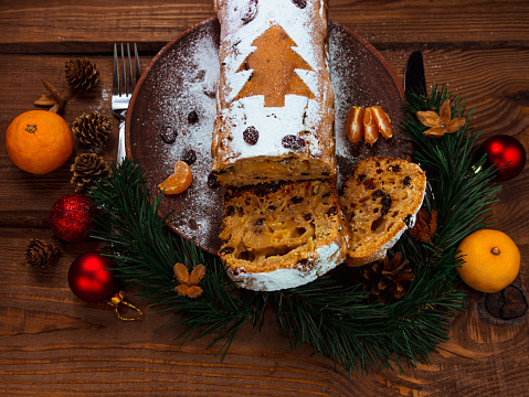 Christmas fruitcake with powdered sugar tangerine cranberry fir tree festive decoration wooden background flat lay. Stollen cake loaf Wholegrain flour dough baking recipe rustic holiday table top view