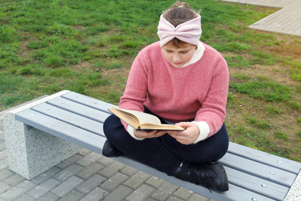 istock Chubby girl sits on bench and thoughtful reads book in park 1355693444