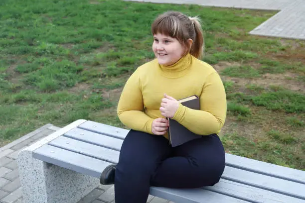 Plump smiling girl sits on bench in park, holds book in her hands and looks away