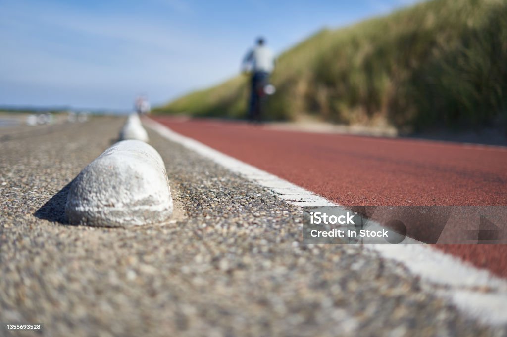 Bicycle path in South Holland. 1 man in dark clothes on bike deep out of focus. Large beach dune with grass. Deep perspective on a day in summer. Netherlands, Schouwen-Duiveland, Brouwersdam. Bicycle Lane Stock Photo