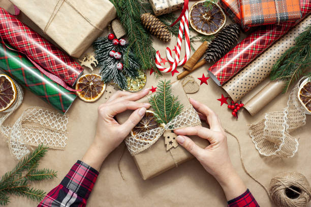 les mains des femmes emballent et décorent un cadeau de noël. la table est recouverte de papier artisanal beige et remplie de matériaux pour la décoration de couleurs rouges et vertes. vue d’en haut - handmade paper paper homemade craft photos et images de collection