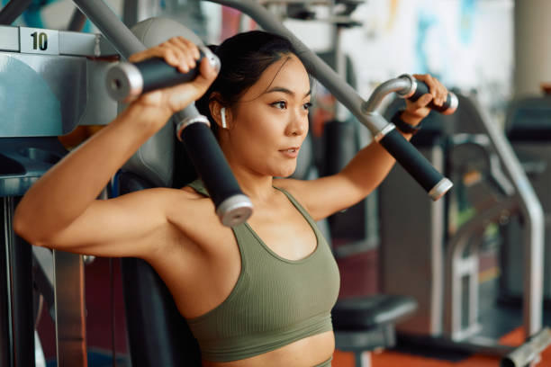 Asian female athlete doing weight exercises for upper body strength during sports training in a gym. Young Asian athletic woman doing chest exercises on a machine while working out in a gym. training equipment stock pictures, royalty-free photos & images