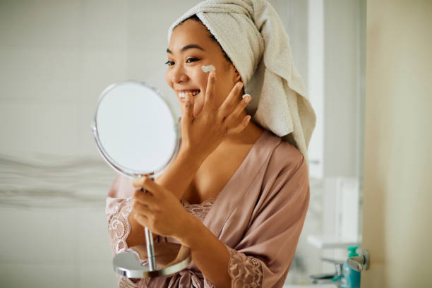 Happy Asian woman applying face cream while looking herself in a mirror in the bathroom. Young Asian woman taking care of her facial skin and applying moisturizer in the bathroom. face cream stock pictures, royalty-free photos & images