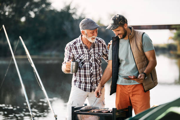 feliz homem maduro falando com seu filho que está grelhando peixe em um churrasco enquanto acampa na natureza. - fishing active seniors family senior adult - fotografias e filmes do acervo