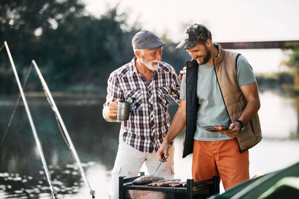 счастливый зрелый мужчина разговаривае�т со своим сыном, который жарит рыбу на барбекю во время кемпинга на природе. - fishing active seniors family senior adult стоковые фото и изображения