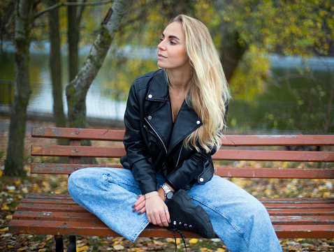 beautiful caucasian blonde female in black leather jacket and jeans is sitting on the branch in the autumn park
