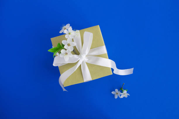 istock Gift box with tied white bow and flowering tree branches on the blue background. Top view. Copy space. 1355703843