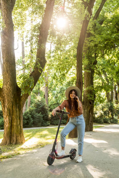 istock A young woman with a hat standing next to her electric scooter while making a phone call. Trees and green colours predominate in the background. 1355705230