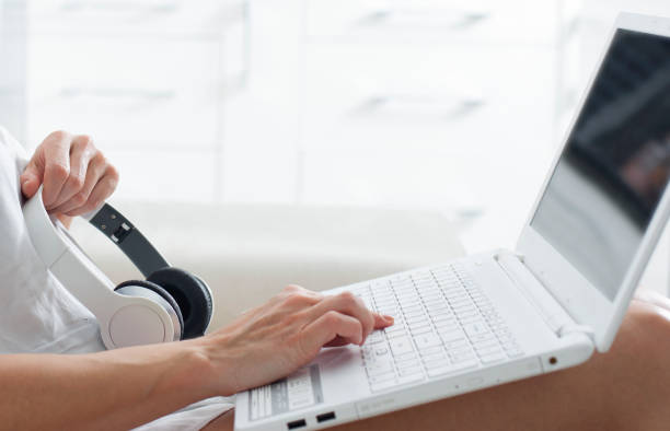 woman sitting on sofa typing on laptop with headphones, dating - typewriter key audio imagens e fotografias de stock