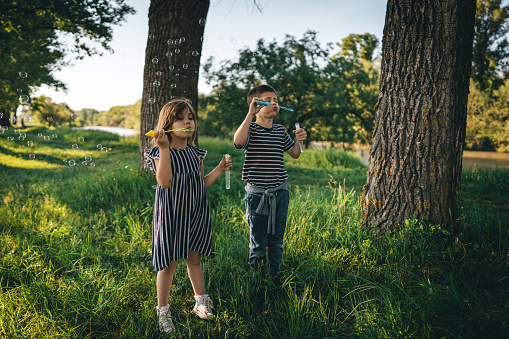 Little brother and sister standing and blowing bubbles with bubble wand in nature