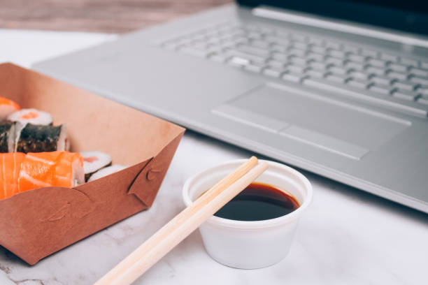 istock Different sushi rolls with salmon in delivery paper box on white marble table background with laptop - futomaki, uramaki and hosomaki. Online order food and delivery food concept 1355713224