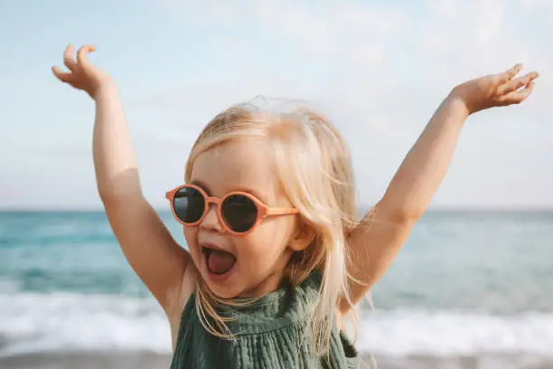 Photo of Funny kid girl playing outdoor surprised emotional child in sunglasses 3 years old baby raised hands family vacations