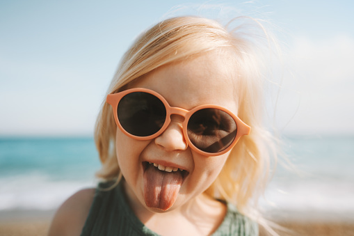 Child sticking out tongue emotional girl in sunglasses 3 years old baby walking on beach family travel lifestyle vacations holidays