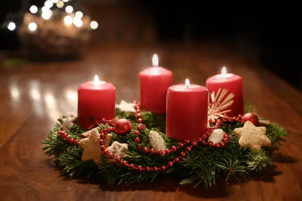 Fourth Advent - decorated Advent wreath from fir branches with red burning candles on a wooden table in the time before Christmas, festive bokeh in the warm dark background, copy space, selected focus