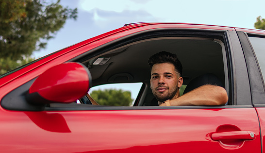 Low angle shot of a young man sitting inside a red car and looking down at the camera through the window of it