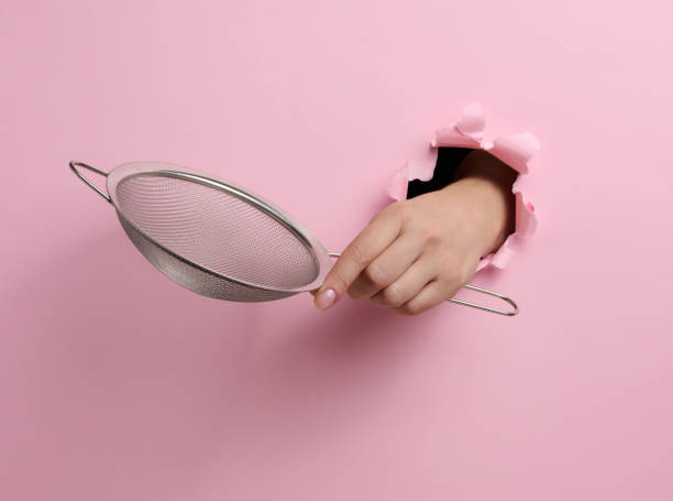 metal sieve for sifting flour in a female hand on a pink background. part of the body sticking out of a torn hole in a paper background - sifting imagens e fotografias de stock