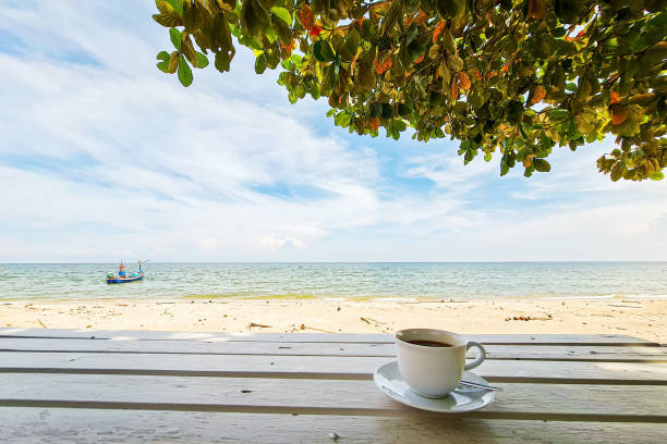 istock Coffee cup mug on white wood table with tree leaf and sea beach ocean background landscape 1355760066