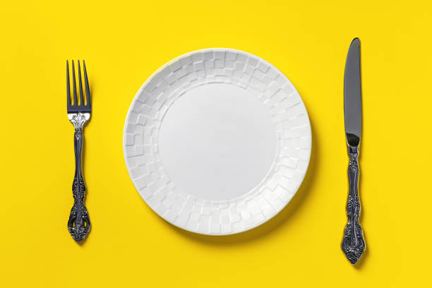 istock Empty white dinner plate, table knife and fork against yellow background. Blank mockup template for unbalanced diets and poor eating habits concepts. 1355784024
