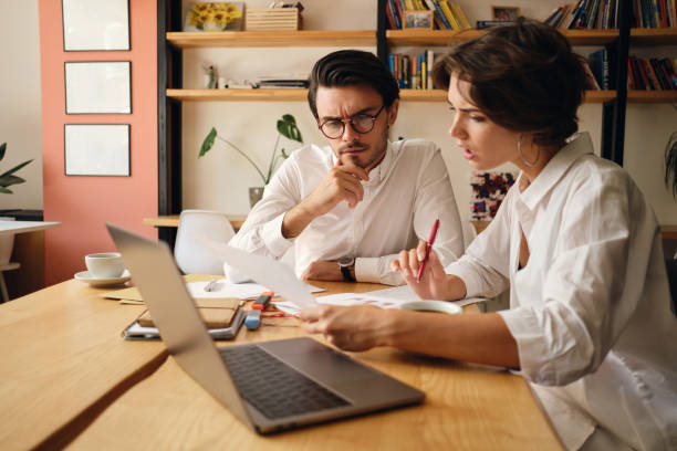 istock Young business colleagues thoughtfully discussing new project sitting at the table with laptop and papers in modern office 1355784137