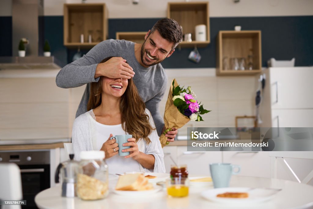 Man holding hand over wife's eyes before he surprises her with bouquet of flowers at home Giving Stock Photo