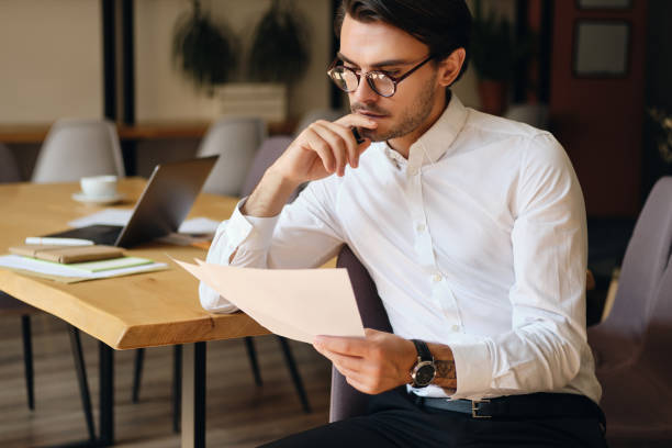 istock Young serious businessman in eyeglasses thoughtfully reading papers working in modern office 1355784793