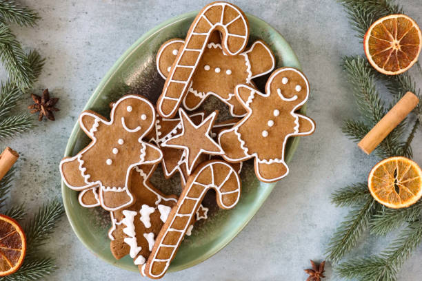 Stock photo showing elevated view of grey background with green, ceramic oval dish of homemade, gingerbread men, stars, candy canes and Christmas tree biscuits decorated with white, royal icing, surrounded by a frame of spruce needles, red berries, cinnamon sticks, star anise and dried citrus orange fruit slices. imagen en primer plano de galletas caseras, con forma de miel con temática navideña heladas con glaseado blanco, real en plato ovalado, rodeado de marco de agujas de abeto, bayas rojas, palitos de canela, anís estrellado y rodajas secas de cítricos ana