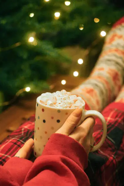 Photo of Image of unrecognisable woman wearing red tartan pyjamas and Christmas themed patterned socks, feet on coffee table, holding spotty, mug of hot chocolate, topped with whipped cream and mini marshmallows, Christmas tree with illuminated fairy lights