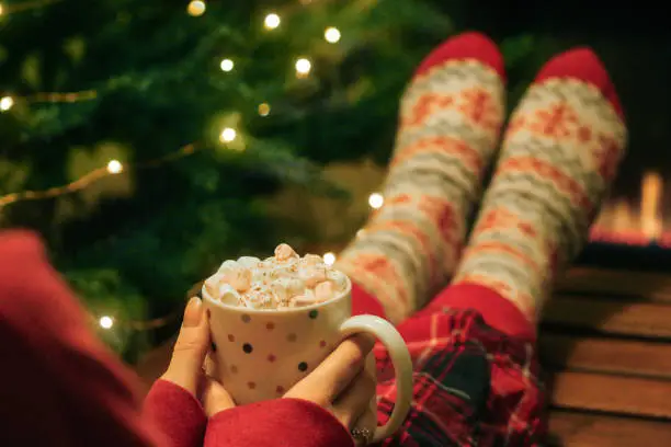 Photo of Image of unrecognisable woman wearing red tartan pyjamas and Christmas themed patterned socks, feet on coffee table, holding spotty, mug of hot chocolate, topped with whipped cream and mini marshmallows, row of gas fire flames in background