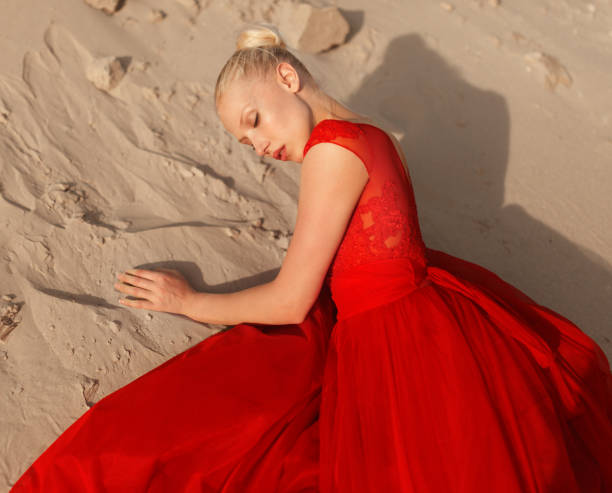 Người mẫu nữ tóc vàng gợi cảm trong chiếc váy đỏ thanh lịch nằm trên cát.