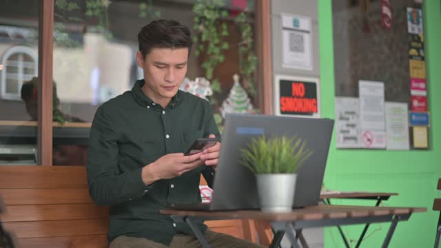 Smiling professional young Asian chinese businessman working using laptop at cafe patio outdoor