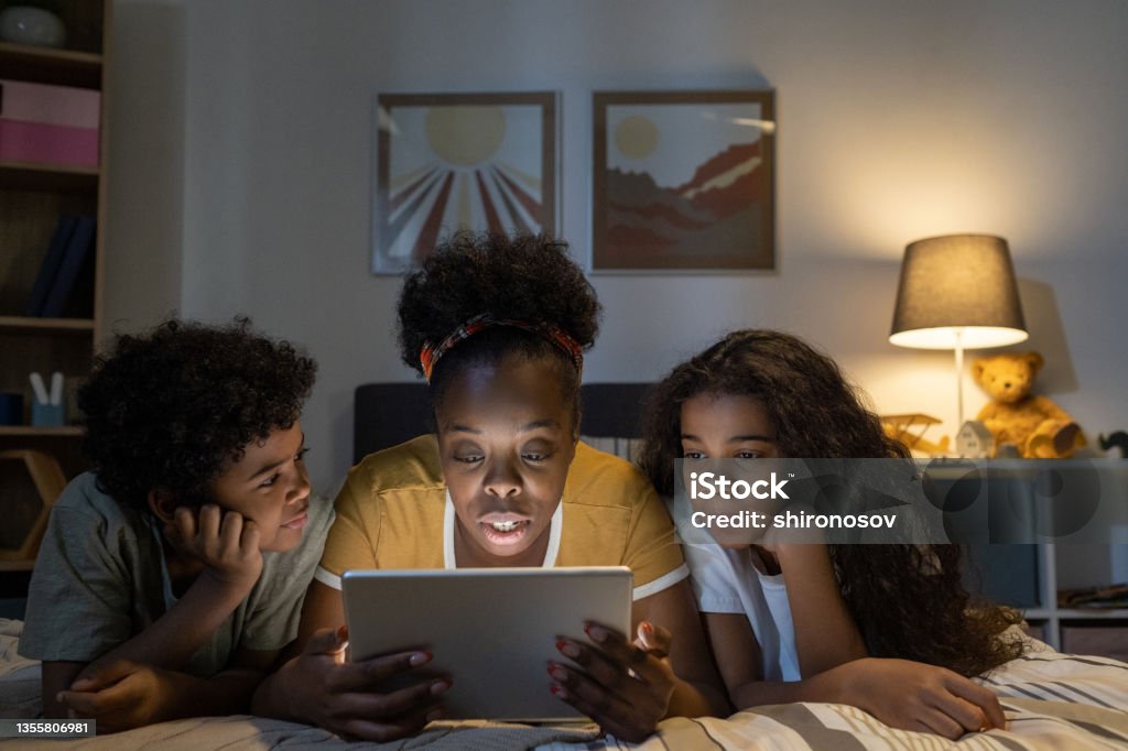 Blackwoman Reading Fairy Tale On Tablet To Kids Middle-aged blackwoman with curly hair lying with kids on bed in nursery room and reading fairy tale on tablet to them 10-11 Years Stock Photo