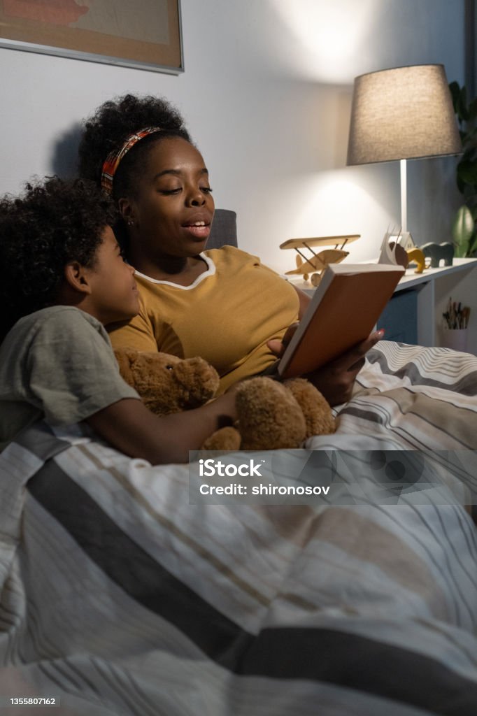 Careful Blackwoman Reading Fairy Tale To Son Careful African American blackwoman lying under duvet in bed and reading fairy tale to son Reading Stock Photo