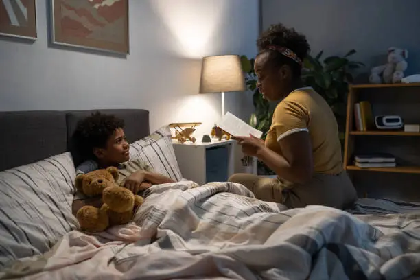 Blackwoman sitting on edge of bed and reading fairy tale to cute son embracing bear toy in bed