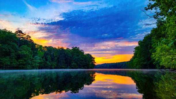 istock Glow of sunrise on partly clouded day over tree lined lake with reflection 1355809837