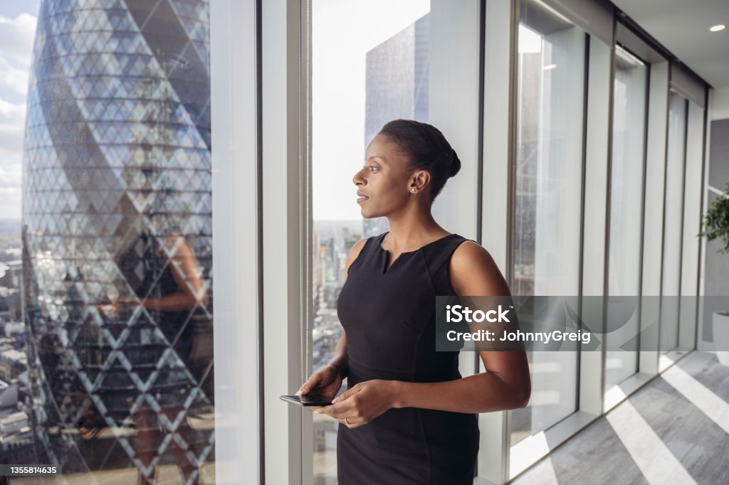 Contemplative British businesswoman standing at window Three-quarter front view of well-dressed Black executive in mid 30s holding smart phone and looking at sunny view of London’s financial district. Office Stock Photo