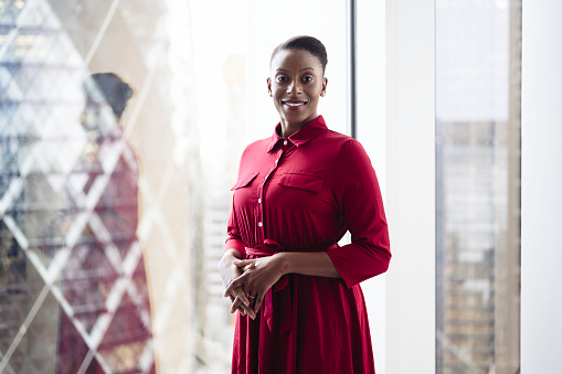 Three-quarter front view of poised Black businesswoman in maroon shirtwaister standing at window smiling at camera with landmark architecture in background.