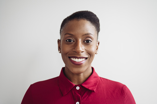 Close-up of executive with hair pulled back wearing maroon shirtwaister and smiling at camera while standing against interior white wall background.