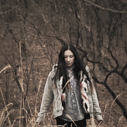 A square photo of a young Japanese woman with long hair in a denim jacket standing in the woods.