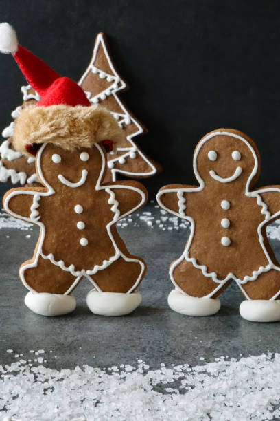 Close-up image of two homemade, gingerbread men skating on pond, one with red Santa hat, in snowy, night Christmas forest scene with tree cookies decorated with white royal icing, black background