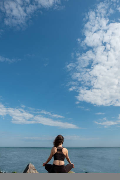 Peaceful meditation. Relaxed woman. Nature harmony. Unrecognizable lady sitting lotus pose on rock stone looking on azure water and blue sky.