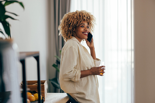 Beautiful mixed race woman standing by her desk talking on the phone. In one hand she is holding her cellphone and in the other she is holding a glass with juice. She is smiling while talking.