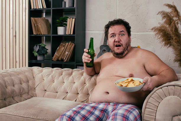 istock Funny fat man drinking beer at home. 1355857933