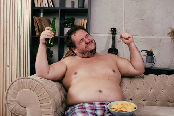 istock Funny fat man drinking beer at home. 1355858156