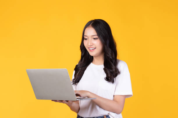 portrait of happy young asian businesswoman, student in white t-shirt working on silver laptop computer isolated on yellow background. - women young adult isolated length imagens e fotografias de stock