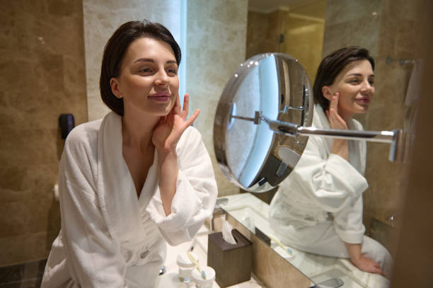 istock Beautiful woman in white bathrobe, smiles looking at her mirror reflection, touching and performing anti-aging smoothing facial massage after applying moisturizer on her face. Skin care concept 1355888578