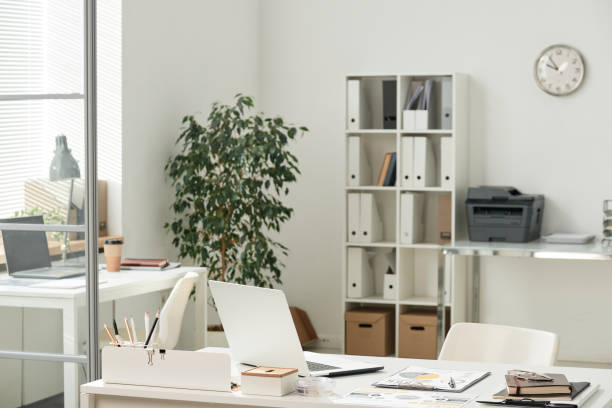 istock Empty modern office workplace with laptops, supplies and papers on tables, shelves with folder and printer in background 1355893093